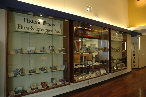 A wall of glass cases containing firefighting photos, placards and memorabilia. A large sign reads "Historic Illinois Fires & Emergencies"